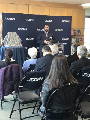 An image capturing a moment of celebration at a UConn event, where Professor Ben Fuller stands at the podium, passionately addressing an audience. The backdrop is adorned with the UConn logo, symbolizing pride in the institution. Seated attendees, including proud family members and university staff, attentively listen, embodying a sense of community and shared achievement. The atmosphere is filled with anticipation and joy, reflecting the significance of the occasion as they gather to honor the graduates and their accomplishments