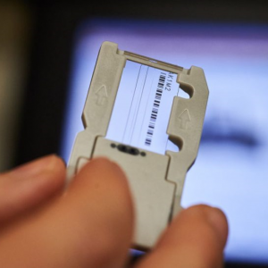 A close-up image of a hand holding a small, transparent slide or card that contains a barcode. The card is positioned in front of a blurred screen displaying a digital image, possibly related to scientific or medical analysis. The background is out of focus, emphasizing the card in the foreground. The overall scene suggests a laboratory or analytical setting where sample identification or data entry is taking place